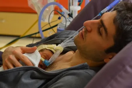 A father holds his premature baby against his chest for skin-to-skin contact, both connected to NICU breathing support.