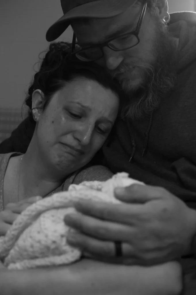 A grieving couple embraces while holding their swaddled baby, with the mother in tears and the father resting his head gently against hers.