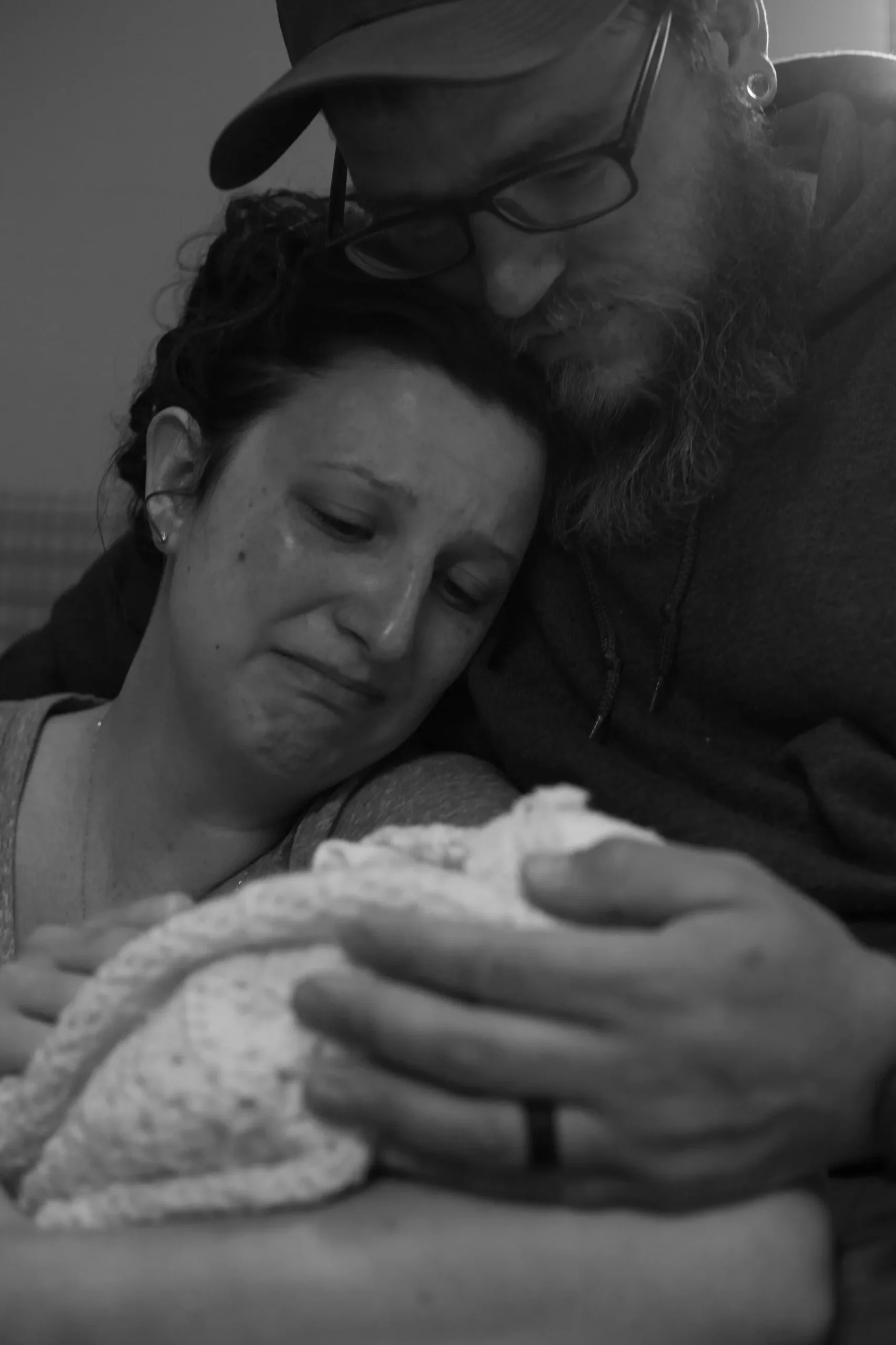 A grieving couple embraces while holding their swaddled baby, with the mother in tears and the father resting his head gently against hers.