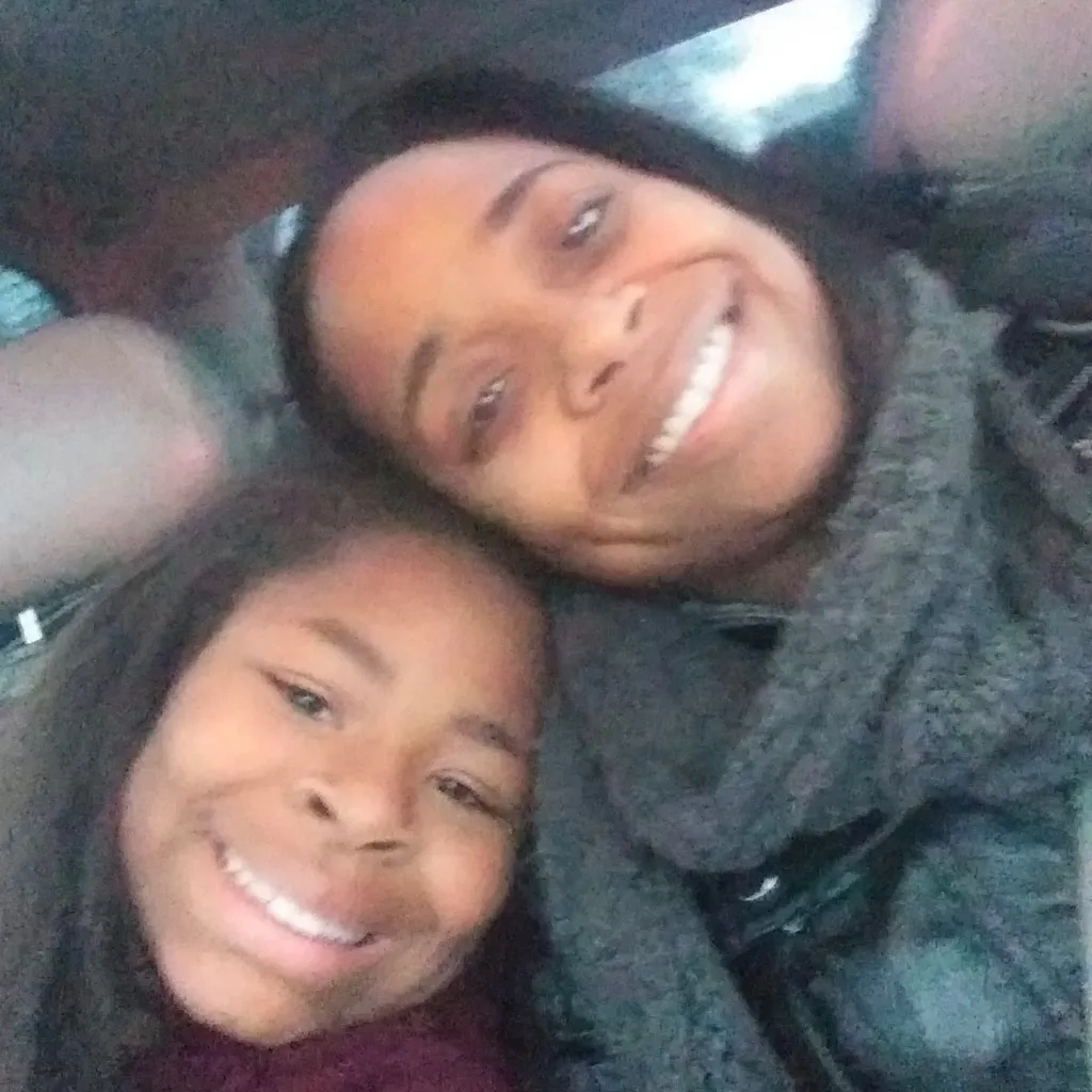 A smiling woman and young girl take a close-up selfie together inside a car, leaning their heads in toward each other.