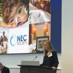 A woman speaks at a podium in front of a large screen displaying the NEC Society logo and images of premature infants, families, and candles.