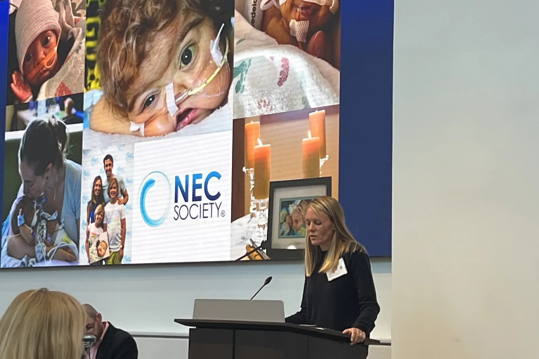 A woman speaks at a podium in front of a large screen displaying the NEC Society logo and images of premature infants, families, and candles.