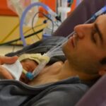 A father holds his premature baby against his chest for skin-to-skin contact, both connected to NICU breathing support.