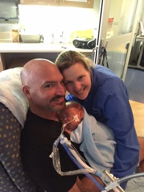 A mother and father smiling while holding their premature baby, who is wrapped in a blanket and connected to medical breathing support in a NICU setting.