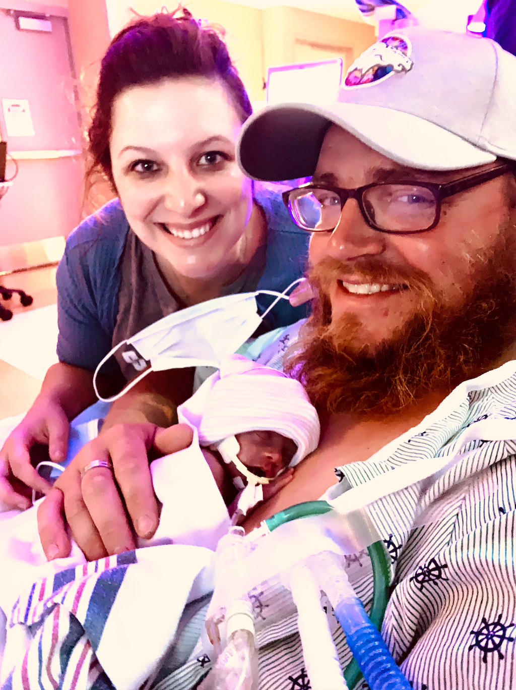 A smiling man and woman pose in a hospital room while holding a premature newborn wearing a tiny hat and receiving breathing support