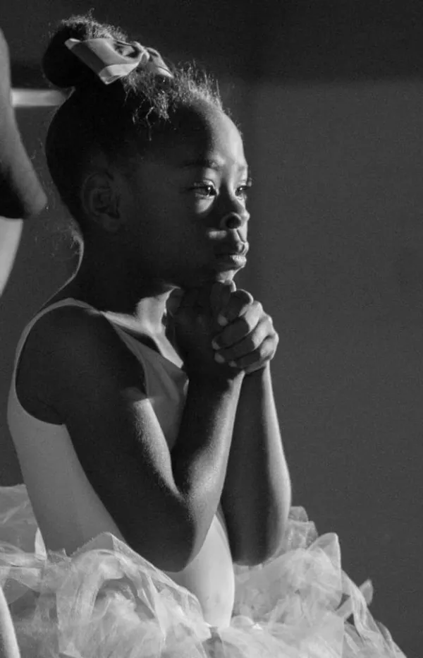 A young girl in a ballet costume stands in profile with hands clasped under her chin, gazing thoughtfully in soft light.