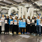 A group of thirteen runners and supporters from the NEC Society pose together in front of large white CIM letters at the California International Marathon expo. They are smiling, wearing athletic gear or NEC Society shirts, and holding posters of babies impacted by NEC, along with a blue sign that reads, “Running 26.2 for the NEC Society: Building a world without NEC.”