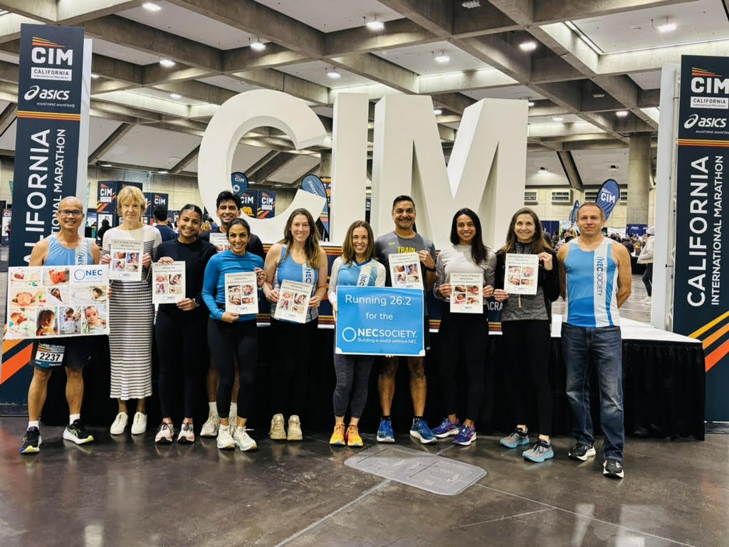 A group of thirteen runners and supporters from the NEC Society pose together in front of large white CIM letters at the California International Marathon expo. They are smiling, wearing athletic gear or NEC Society shirts, and holding posters of babies impacted by NEC, along with a blue sign that reads, “Running 26.2 for the NEC Society: Building a world without NEC.”