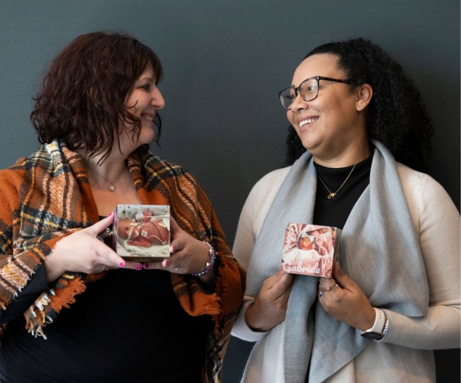 Cassey and Tabitha holding pictures of their daughters who tragically passed away from NEC, smiling at each other